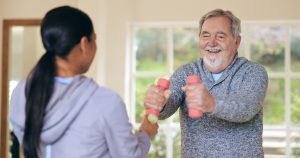 Woman, elderly man and workout with dumbbells, health and happy for progress in nursing home. Personal trainer, physiotherapist and training for muscle, rehabilitation and exercise in retirement